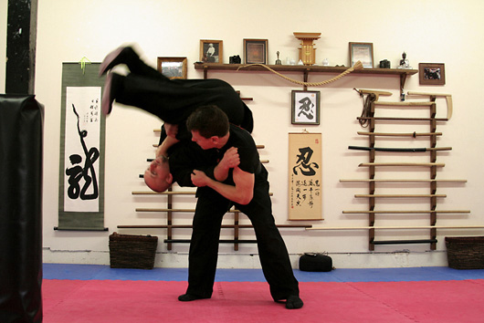A throw being executed in the Black Snow dojo: black gi, blue mats, the weapon rack and hanging scrolls behind.