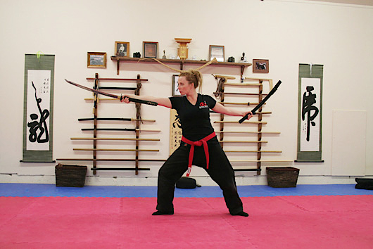 A student in a weapons stance with a bokken in each hand, red belt, in front of the weapon rack and scrolls.