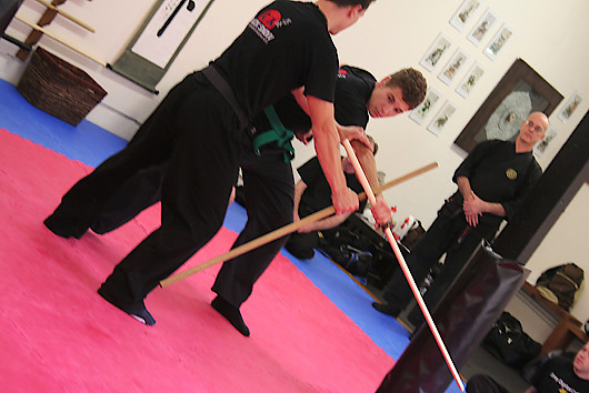Three adults practising bo staff work on the pink mat.