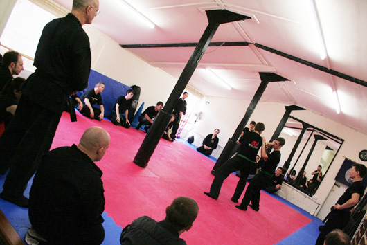 A wide view of the dojo interior: mirror wall, mats and students seated.